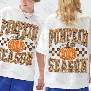 May include: A white short-sleeved t-shirt featuring a graphic print of an orange pumpkin centered between the words PUMPKIN and SEASON in large block letters. The text has a brown and orange autumn leaf pattern with a brown checkered border.