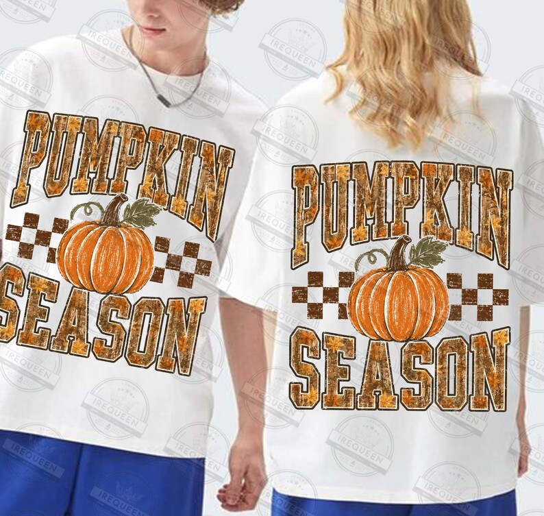 May include: A white short-sleeved t-shirt featuring a graphic print of an orange pumpkin centered between the words PUMPKIN and SEASON in large block letters. The text has a brown and orange autumn leaf pattern with a brown checkered border.