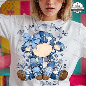 Könnte beinhalten: Weißes Langarmshirt mit einem Patchwork-Lamm-Design und einer blauen Schleife. Der Text "The Lord is my Shepherd" und "Psalm 23:1" sind ebenfalls sichtbar. Das Lamm besteht aus verschiedenen Blautönen.