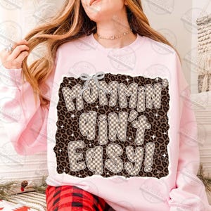 以下が含まれることがあります： ヒョウ柄の背景に、茶色と白のチェック柄で「Mommin' Ain't Easy!」と書かれた薄いピンク色のスウェットシャツ。テキストの上部にはシルバーのリボンが付いています。