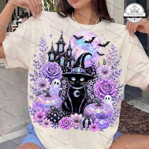 May include: A cream-colored t-shirt featuring a graphic print of a black cat wearing a witch hat, surrounded by purple roses, pumpkins, ghosts, a haunted house, and a large moon with bats. The design includes a crescent moon charm on the cat's collar.