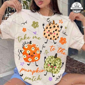 Puede incluir: Una camiseta de manga corta de color crema con un estampado gráfico de tres calabazas de dibujos animados con caras, brazos y piernas. El texto "take me to the pumpkin patch" aparece en letras naranjas y verdes. Pequeñas flores y estrellas.