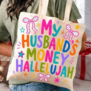Pu&ograve; includere: Una borsa in tela color crema con il testo colorato "My Husband's Money Hallelujah" in un carattere giocoso. Il design &egrave; decorato con fiocchi a pois rosa e bianchi e varie piccole stelle nei colori arancione, rosa e blu.