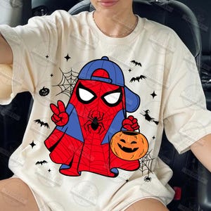 Pode incluir: Camiseta creme com um design tem&aacute;tico de Halloween. O design apresenta um fantasma vestido de Homem-Aranha, usando um bon&eacute; azul e segurando uma lanterna de ab&oacute;bora. Morcegos pretos, teias de aranha e estrelas completam o visual festivo.