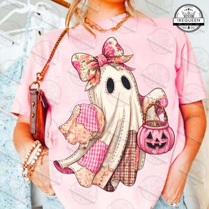 May include: A pink t-shirt featuring a whimsical ghost design. The ghost wears a floral bow and holds a pink pumpkin basket. The ghost's patchwork design includes pink and brown fabrics, perfect for Halloween.
