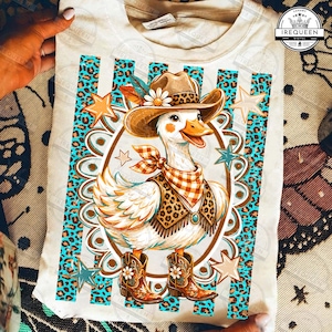 May include: A cream-coloured t-shirt featuring a graphic of a white duck wearing a brown cowboy hat with a leopard print band, a checkered bandana, a leopard print vest, and brown cowboy boots, surrounded by turquoise leopard print stripes.