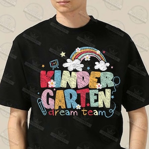 May include: Black t-shirt with a colorful graphic design. The design features the words "Kindergarten dream team" in various colors, a rainbow, clouds, stars, and flowers. A pencil and a math symbol are also included.