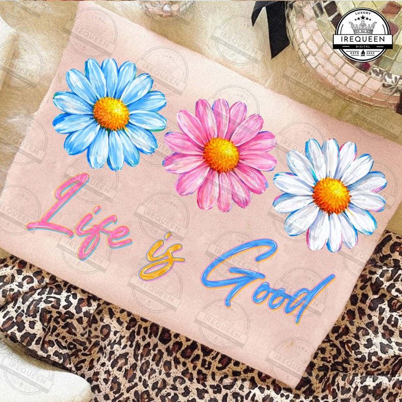Pu&ograve; includere: Una maglietta rosa chiaro con tre grandi margherite blu, rosa e bianche con centri gialli. Le parole "Life is Good" sono scritte in corsivo in rosa, giallo e blu. La maglietta &egrave; appoggiata su un tessuto con stampa leopardata.