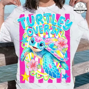 May include: White t-shirt featuring a colorful graphic of a cartoon turtle with the text "TURTLEY OVER IT". The turtle is blue, pink, and yellow, with hibiscus flowers and stars. The background has pink stripes.