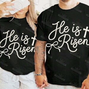 May include: Black t-shirts with the text "He is Risen" in a white, cursive font. The design includes a cross and a bow. The shirts are likely for Easter or religious events.