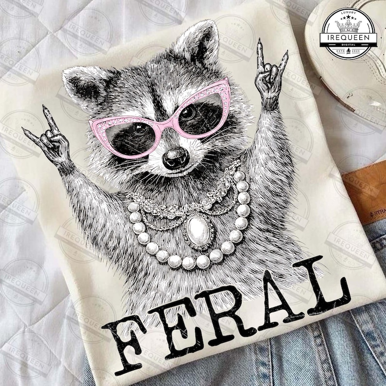 Puede incluir: Una camiseta color crema con una ilustraci&oacute;n detallada de un mapache con gafas de sol rosas y un collar de perlas. El mapache est&aacute; haciendo un gesto de rock and roll con la mano. La palabra "FERAL" est&aacute; impresa en letras negras en negrita.
