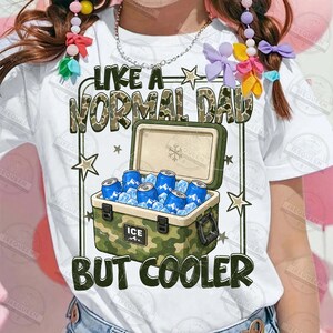 May include: White t-shirt with the text "LIKE A NORMAL DAD BUT COOLER" in green and camouflage print. The graphic features an open camouflage cooler filled with blue cans and ice cubes. Stars and lines frame the text and cooler.