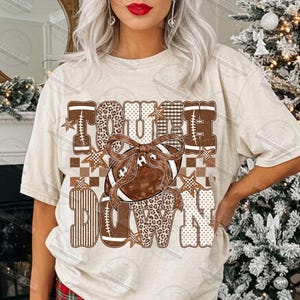 Peut inclure: Un t-shirt couleur cr&egrave;me avec un motif graphique portant le texte TOUCHDOWN dans des tons marron et blanc, incluant des imprim&eacute;s l&eacute;opard, &agrave; carreaux et ray&eacute;s. Un ballon de football marron avec un grand n&oelig;ud est centr&eacute; avec des &eacute;toiles.