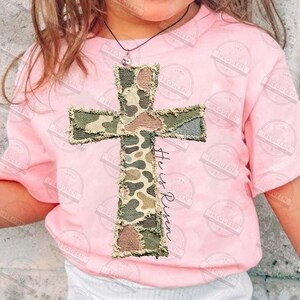 May include: A light pink t-shirt featuring a camouflage cross applique with frayed edges. The text "He is Risen" is printed vertically on the cross. The shirt is worn by a person.
