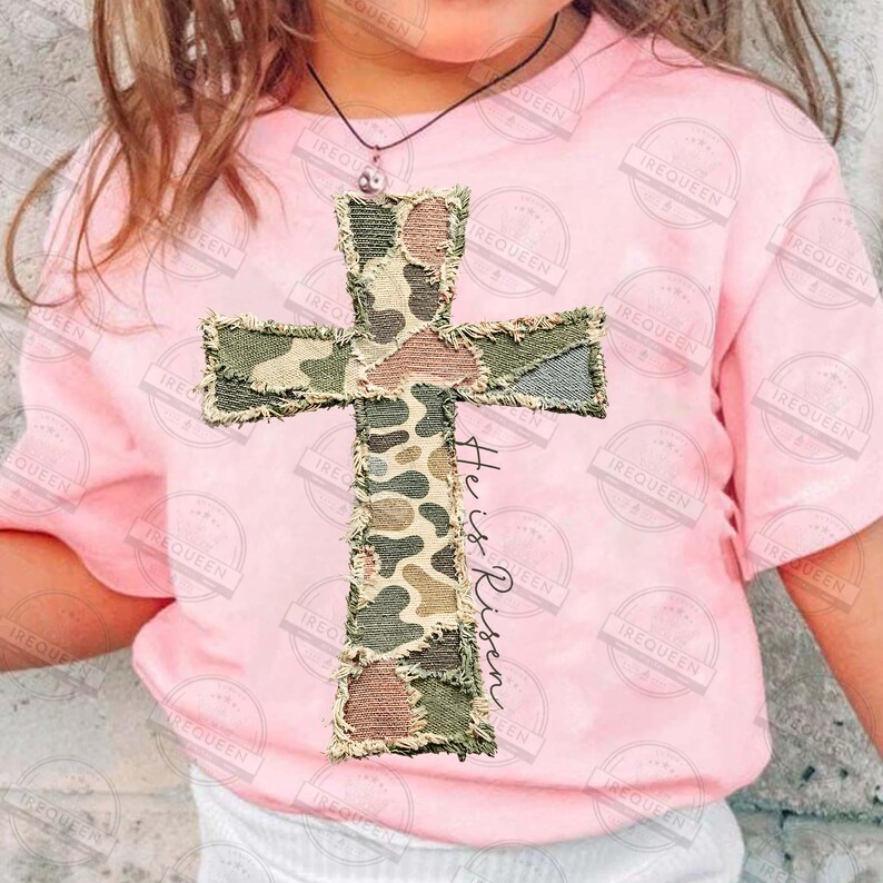 May include: A light pink t-shirt featuring a camouflage cross applique with frayed edges. The text "He is Risen" is printed vertically on the cross. The shirt is worn by a person.
