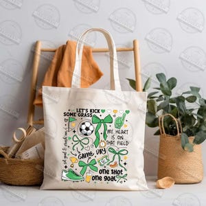 May include: White canvas tote bag with a green, yellow, and white design featuring a soccer ball, bows, and the text "Let's Kick Some Grass", "My Heart Is On That Field", "Game Day", and "One Shot One Goal".