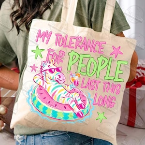 Puede incluir: Bolso tote beige con el texto "MY TOLERANCE FOR PEOPLE LAST THIS LONG" en rosa y verde. Presenta un cebra con gafas de sol flotando en una boya con forma de sand&iacute;a.