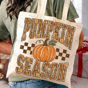 May include: A cream-colored canvas tote bag featuring a large orange pumpkin graphic in the center. The text PUMPKIN SEASON is printed in bold, block letters with a brown and orange autumn leaf pattern. A brown checkered pattern sits behind the pumpkin.
