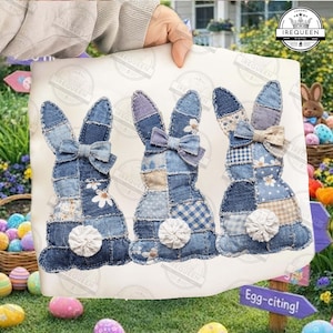 Op de afbeelding: Een decoratief stoffen paneel met drie denim patchwork konijnen, elk met een strik en een pluizige witte staart. De konijnen zijn in tinten blauw en wit, met bloemen- en vichy-accenten. Het paneel wordt door een persoon omhoog gehouden.