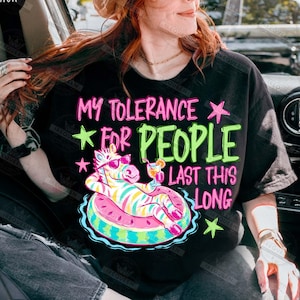 Puede incluir: Camiseta negra con el texto "MY TOLERANCE FOR PEOPLE LAST THIS LONG" en rosa y verde. El gr&aacute;fico presenta una cebra con gafas de sol flotando en un flotador con forma de sand&iacute;a con un c&oacute;ctel.