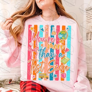 Może przedstawiać: Jasnor&oacute;żowa bluza z kolorowymi pionowymi paskami i napisem "You're more than a Test Score". Projekt zawiera globus, szkolny autobus, jabłko i inne grafiki związane ze szkołą.