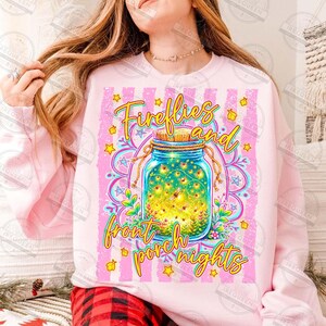 May include: Pink sweatshirt with a graphic of a mason jar filled with fireflies, the text "Fireflies and front porch nights" in yellow script. The design includes stars and a floral mandala.