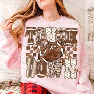 Peut inclure: Un sweat-shirt &agrave; manches longues rose clair avec le texte TOUCH DOWN en grandes lettres majuscules. Les lettres pr&eacute;sentent des motifs marron, blanc et beige, notamment des imprim&eacute;s l&eacute;opard, carreaux et rayures. Un ballon de football marron avec un n&oelig;ud est au centre.