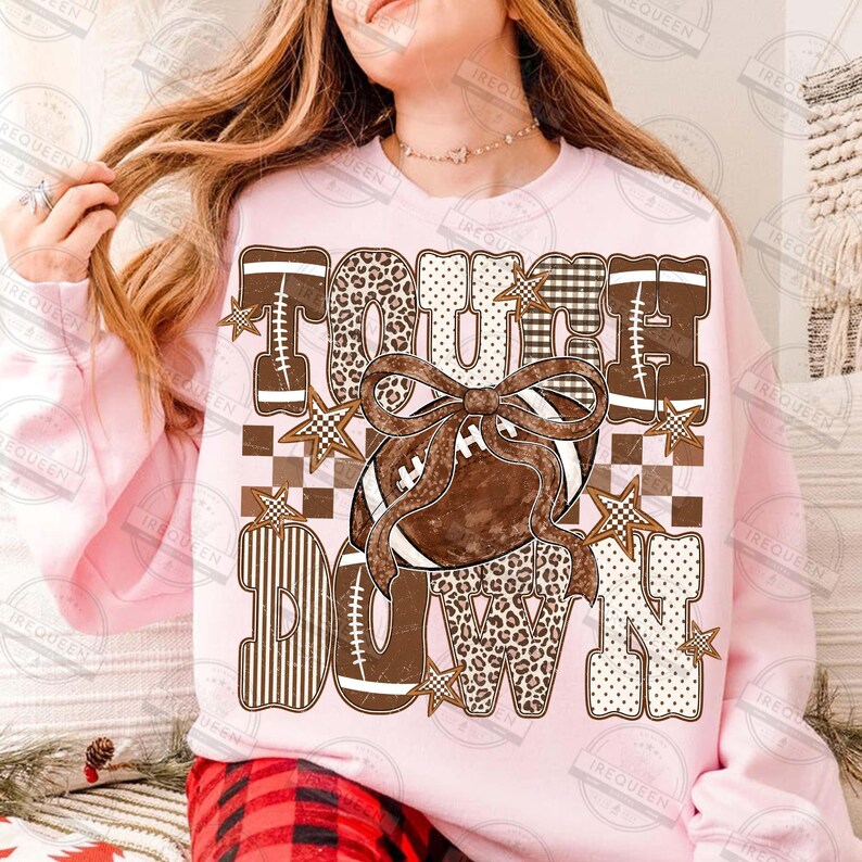 Peut inclure: Un sweat-shirt &agrave; manches longues rose clair avec le texte TOUCH DOWN en grandes lettres majuscules. Les lettres pr&eacute;sentent des motifs marron, blanc et beige, notamment des imprim&eacute;s l&eacute;opard, carreaux et rayures. Un ballon de football marron avec un n&oelig;ud est au centre.