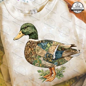 Könnte beinhalten: Ein cremefarbenes Sweatshirt mit einer detaillierten Grafik einer Ente. Die Ente hat einen grünen Kopf, einen gelben Schnabel und einen Körper, der aus Patchwork-Mustern mit floralen und botanischen Designs besteht. Die Ente steht auf grünen Blättern.