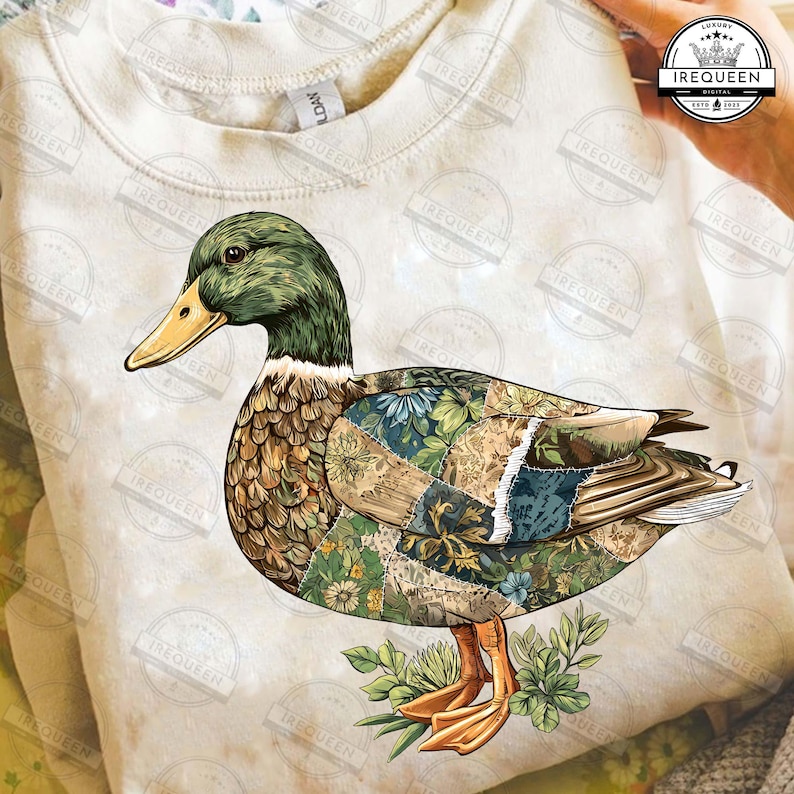 May include: A cream-colored sweatshirt featuring a detailed graphic of a duck. The duck has a green head, a yellow beak, and a body composed of patchwork patterns with floral and botanical designs. The duck is standing on green leaves.