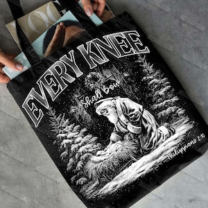 May include: Black tote bag featuring a black and white illustration of Santa kneeling before baby Jesus. The text "EVERY KNEE Shall Bow" is displayed above the image, with "Philippians 2:10" below.
