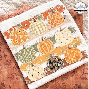 May include: A white fabric square featuring a graphic print of nine pumpkins in various patterns and colors, including orange, green, black, and cream. The pumpkins are arranged over horizontal brushstroke stripes with small stars and hearts.
