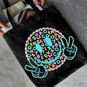May include: Black tote bag featuring a colorful smiley face design. The smiley face is composed of various letters and symbols, with peace signs on each side. The bag is made of a durable material and is suitable for carrying books or groceries.