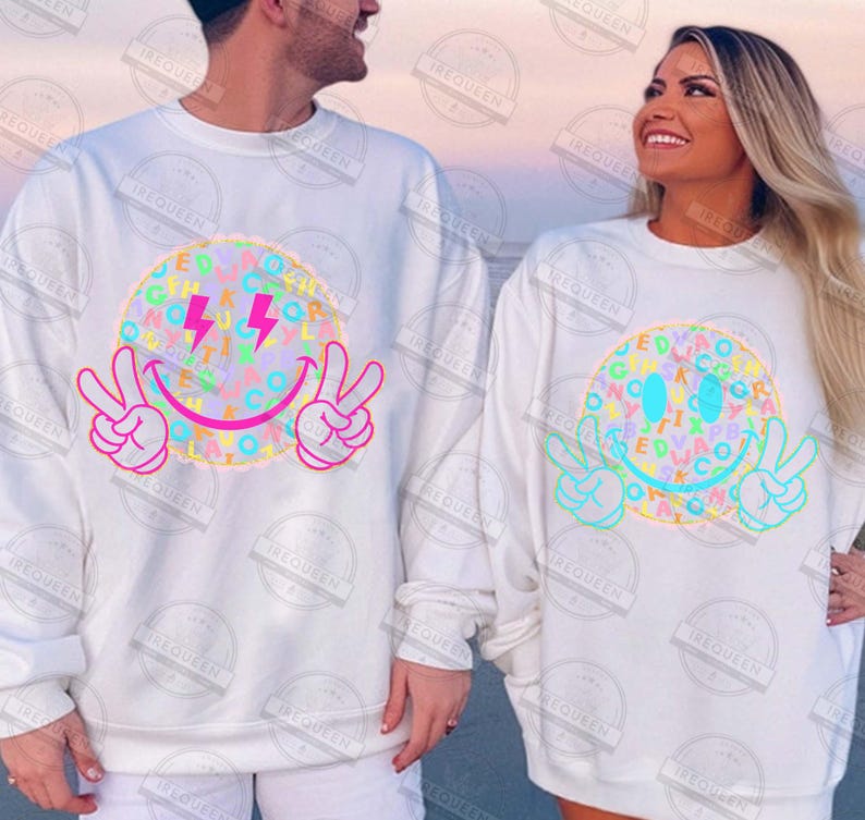 May include: White sweatshirts featuring a colorful smiley face design. The design includes a smiley face with peace signs, lightning bolts, and letters. The sweatshirts are long-sleeved and suitable for casual wear.