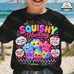 Może przedstawiać: Czarny t-shirt z kolorową grafiką zawierającą napis THE YEAR OF SQUISHY HUNTING, Life is better with squishies, Just a girl who loves squishies, Squish Squad, In My Squishies Era, Madison oraz 2026, otoczony tęczowymi ikonami.