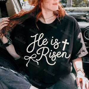 May include: Black t-shirt with the text "He is Risen" in a white, pearl-like font. The design includes a cross and a bow. The shirt is a casual style, suitable for everyday wear.