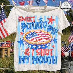 May include: Cream-colored t-shirt with a patriotic design. The graphic features a sweet potato pie decorated with an American flag, surrounded by stars and the text "Sweet Potato Pie & I Shut My Mouth."