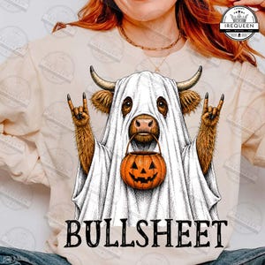 Può includere: Una felpa beige a maniche lunghe con la grafica di una mucca highland marrone vestita da fantasma bianco. La mucca fa un gesto rock-and-roll con la mano e tiene in mano un secchiello a forma di zucca arancione. Il testo BULLSHEET è stampato sotto.