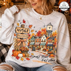 May include: A white crewneck sweatshirt featuring a colorful graphic of a small town street during autumn. The design includes a wooden signpost labeled 47567 Petersburg, a water tower, storefronts, pumpkins, and the text Small Town Fall Vibes.