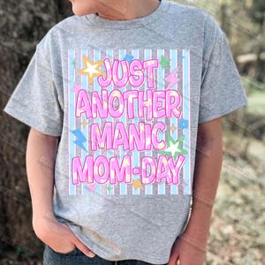 May include: A gray t-shirt with the text "JUST ANOTHER MANIC MOM-DAY" in pink bubble letters. The text is set against a light blue and white striped background, with star, lightning bolt, and flower graphics.