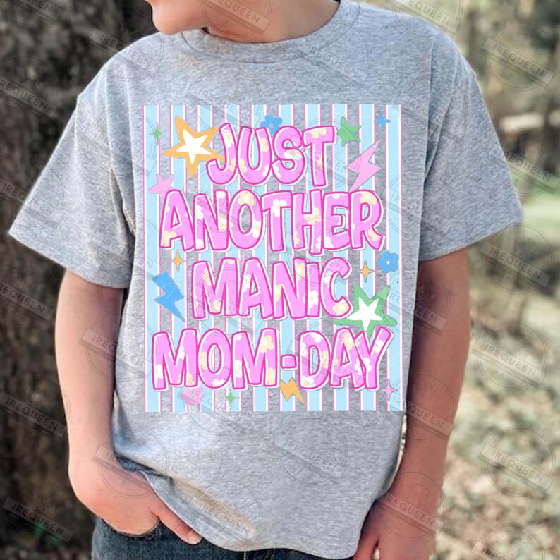 May include: A gray t-shirt with the text "JUST ANOTHER MANIC MOM-DAY" in pink bubble letters. The text is set against a light blue and white striped background, with star, lightning bolt, and flower graphics.