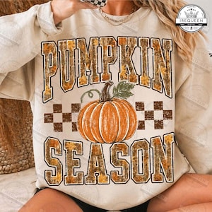 May include: A beige crewneck sweatshirt featuring the text PUMPKIN SEASON in a block font with a camouflage and leaf pattern. A central orange pumpkin illustration sits between the words, accented by a brown and white checkered border design.