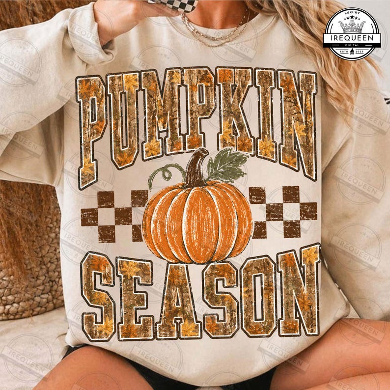 May include: A beige crewneck sweatshirt featuring the text PUMPKIN SEASON in a block font with a camouflage and leaf pattern. A central orange pumpkin illustration sits between the words, accented by a brown and white checkered border design.