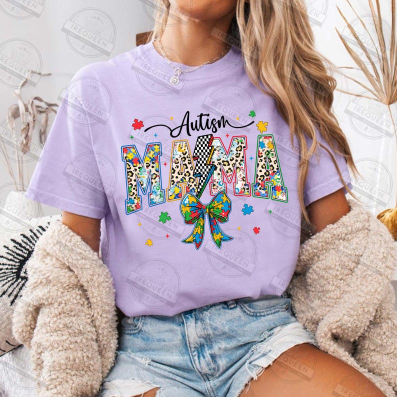 May include: Purple t-shirt with the text "Autism Mama" in colorful puzzle piece letters with a leopard print background. The letters are decorated with a lightning bolt and a rainbow ribbon.