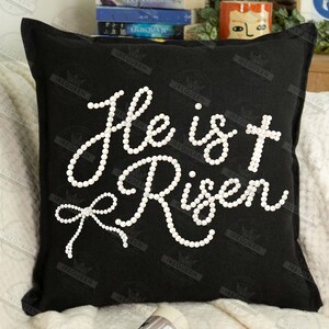 May include: Black square pillow with the words "He is Risen" spelled out in white pearl-like beads. The pillow has a bow design at the bottom of the text.