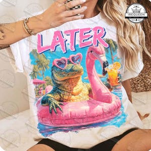 Peut inclure: T-shirt blanc avec un motif représentant un alligator de dessin animé portant des lunettes de soleil en forme de cœur et un flotteur de flamant rose. Le mot "LATER" est imprimé en rose. Scène de plage tropicale.