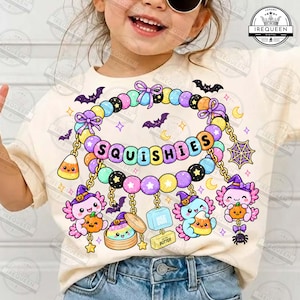May include: A cream-colored t-shirt featuring a graphic of a beaded bracelet with the word SQUISHIES in the center. The design includes cartoon axolotls wearing witch hats, candy corn, a spider web, bats, and small stars in pastel colors.