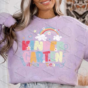 May include: A light purple sweatshirt with the words "KINDERGARTEN" in colorful block letters, decorated with a rainbow, clouds, and flowers. The word "team" is also visible. The sweatshirt is a comfortable, casual style.