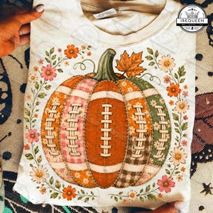 Può includere: Una t-shirt color crema con una grafica centrale raffigurante una zucca disegnata con lacci in stile pallone da football. La zucca è composta da vari segmenti a motivi geometrici in arancione, rosa e verde, circondata da fiori autunnali.