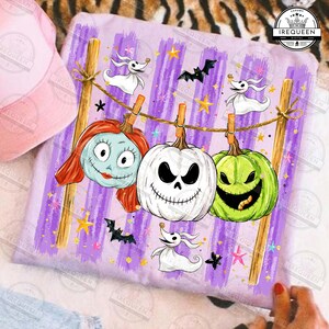 May include: A light purple t-shirt featuring a graphic print of three jack-o'-lanterns and two ghost characters hanging from a clothesline. The design includes small bats, stars, and sparkles against a background of vertical purple brush strokes.